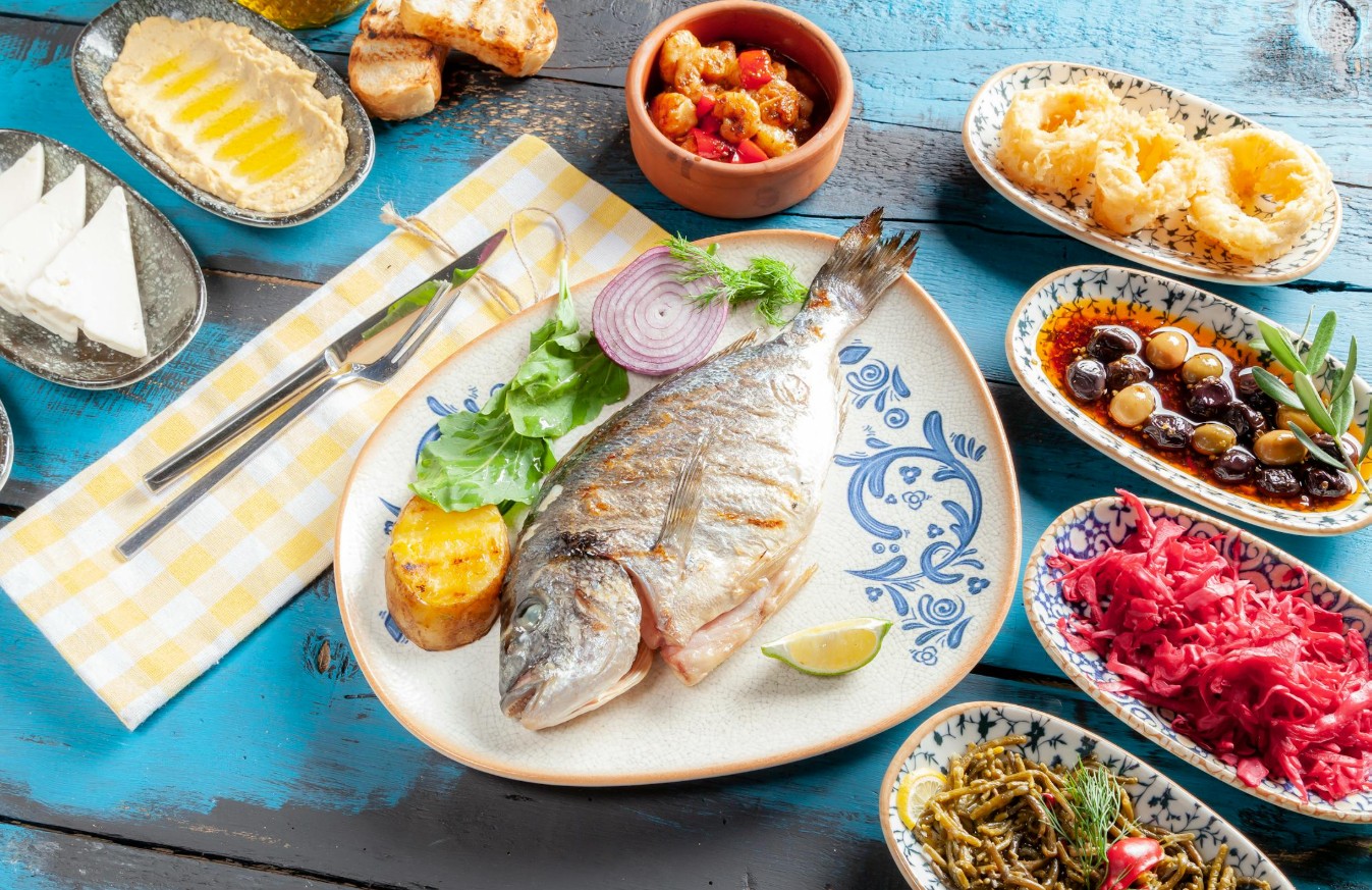 Gegrilde dorade met mediterrane bijgerechten zoals hummus, olijven, feta en gegrilde groenten op een blauw houten tafel