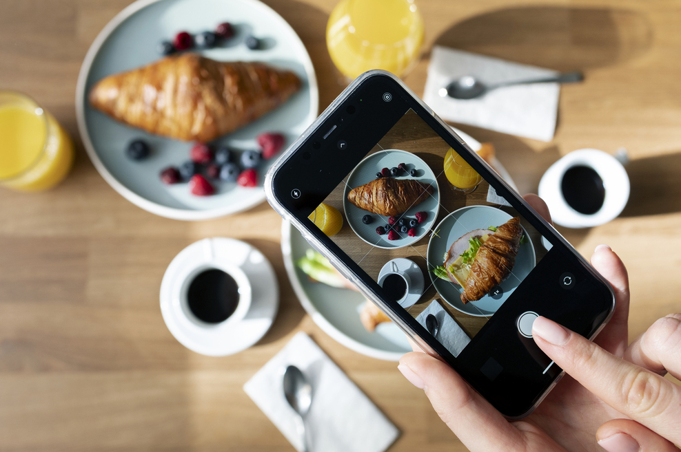 Croissant op ontbijttafel met koffie, sinaasappelsap en smartphone die een foodfoto maakt in de horeca.